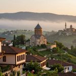 A breathtaking panoramic view of historical Trabzon, showcasing its lush green landscapes and misty horizons. The foreground features traditional Ottoman architecture, with wooden balconies adorned with colorful flowers. In the middle ground, ancient stone churches and fortresses rise majestically, surrounded by verdant hills. The background reveals dramatic mist-covered mountains under a soft golden light of dawn, creating an enchanting atmosphere. Capture the image from a slightly elevated angle to emphasize depth, with the sun gently illuminating the scene, enhancing the rich textures of the buildings and the vibrant greenery. The mood should convey a sense of timeless beauty and tranquility, inviting viewers to immerse themselves in this unforgettable landscape.