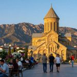 A picturesque view of Kutaisi, the cultural capital of Georgia, showcasing its architectural elegance and natural beauty. In the foreground, a lively plaza with locals dressed in modest casual clothing, enjoying coffee at outdoor cafes. The middle ground features the stunning Bagrati Cathedral, its ancient stone structure bathed in warm golden sunlight, surrounded by vibrant greenery and blooming flowers. In the background, the rugged Caucasus Mountains rise majestically against a clear blue sky, adding a sense of grandeur. The scene conveys a warm, inviting atmosphere, highlighting the city's role as a gateway to Western charm. Capture this moment during the golden hour with soft, diffused lighting to enhance the colors and create a serene mood.