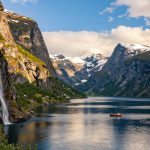 A breathtaking scene of the Norwegian fjords, showcasing steep cliffs rising dramatically from deep blue waters. In the foreground, vibrant green foliage clings to the rocky edges, while waterfalls cascade down into the fjord below. In the middle ground, a small wooden boat sails peacefully, its reflection shimmering in the water. The background features majestic snow-capped mountains rising under a clear blue sky with fluffy white clouds scattered throughout. The lighting is warm and soft, suggesting late afternoon, casting gentle shadows that enhance the textures of the landscape. The overall atmosphere is serene and awe-inspiring, capturing the legendary beauty and adventurous spirit of Norway's natural wonders.